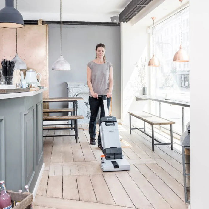 Woman operating Nilfisk SC100 upright scrubber dryer in a restaurant, demonstrating professional floor cleaning performance.